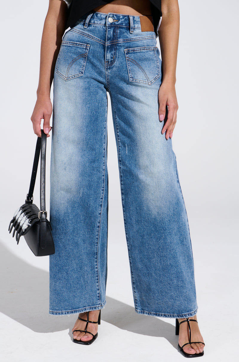 A woman in REVERSE REVERSE MEDIUM WASH JEANS and a black top carries a chic black handbag with chain detail. Her look is completed with stylish black heeled sandals, exuding effortless elegance.