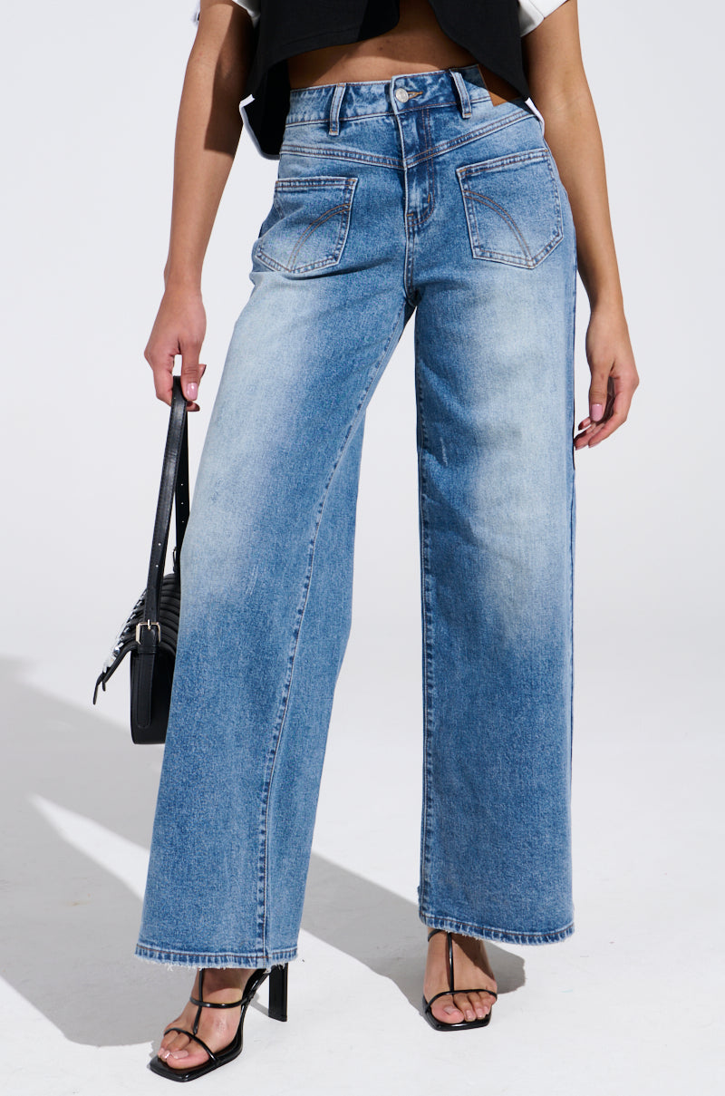 A person wears REVERSE REVERSE MEDIUM WASH JEANS, featuring a high-rise fit and wide-leg style. They pair it with a black cropped vest, accessorize with a black handbag, and finish the outfit with sleek black strappy heels, all posed against a plain white backdrop.