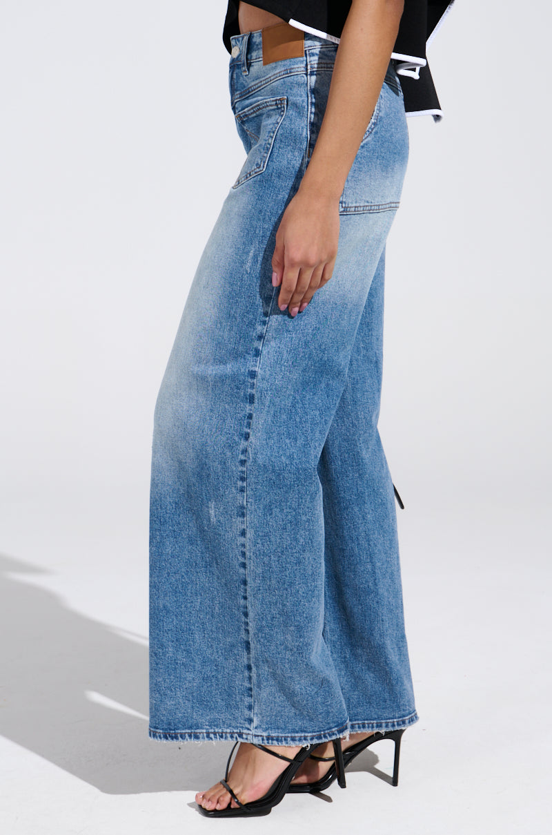 A person is shown from the side, highlighting their wide-leg silhouette and black open-toe heels. The REVERSE REVERSE MEDIUM WASH JEANS have a high-rise fit with pockets, complemented by a faded blue color against a plain white background.