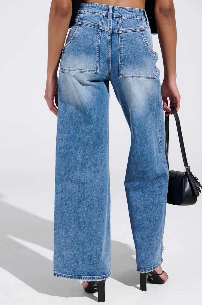 Dressed in REVERSE REVERSE MEDIUM WASH JEANS, featuring a high-rise fit and wide-leg style with subtle fading on the back pockets and legs, a person confidently holds a small black handbag against a neutral background, epitomizing effortless style.