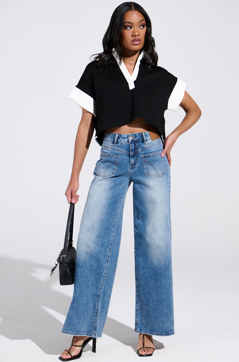 A woman exudes confidence in REVERSE REVERSE MEDIUM WASH JEANS, featuring a high-rise fit and wide-leg style. She pairs them with a black cropped top accented in white, finishing her look with black heels and a bag. Her wavy hair flows against the plain white backdrop.