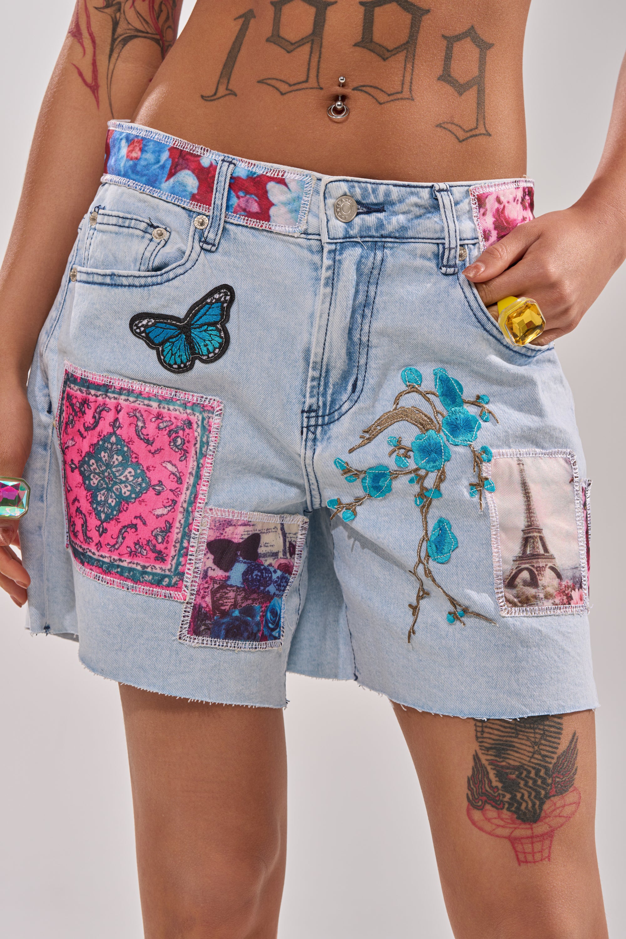 Wearing GET THE PICTURE PATCHWORK DENIM SHORTS featuring a high-rise waist, raw-cut hem, and vibrant patches—blue butterfly, blue flowers, pink bandana print, and Eiffel Tower. Visible tattoos on body and hand add edge. 0