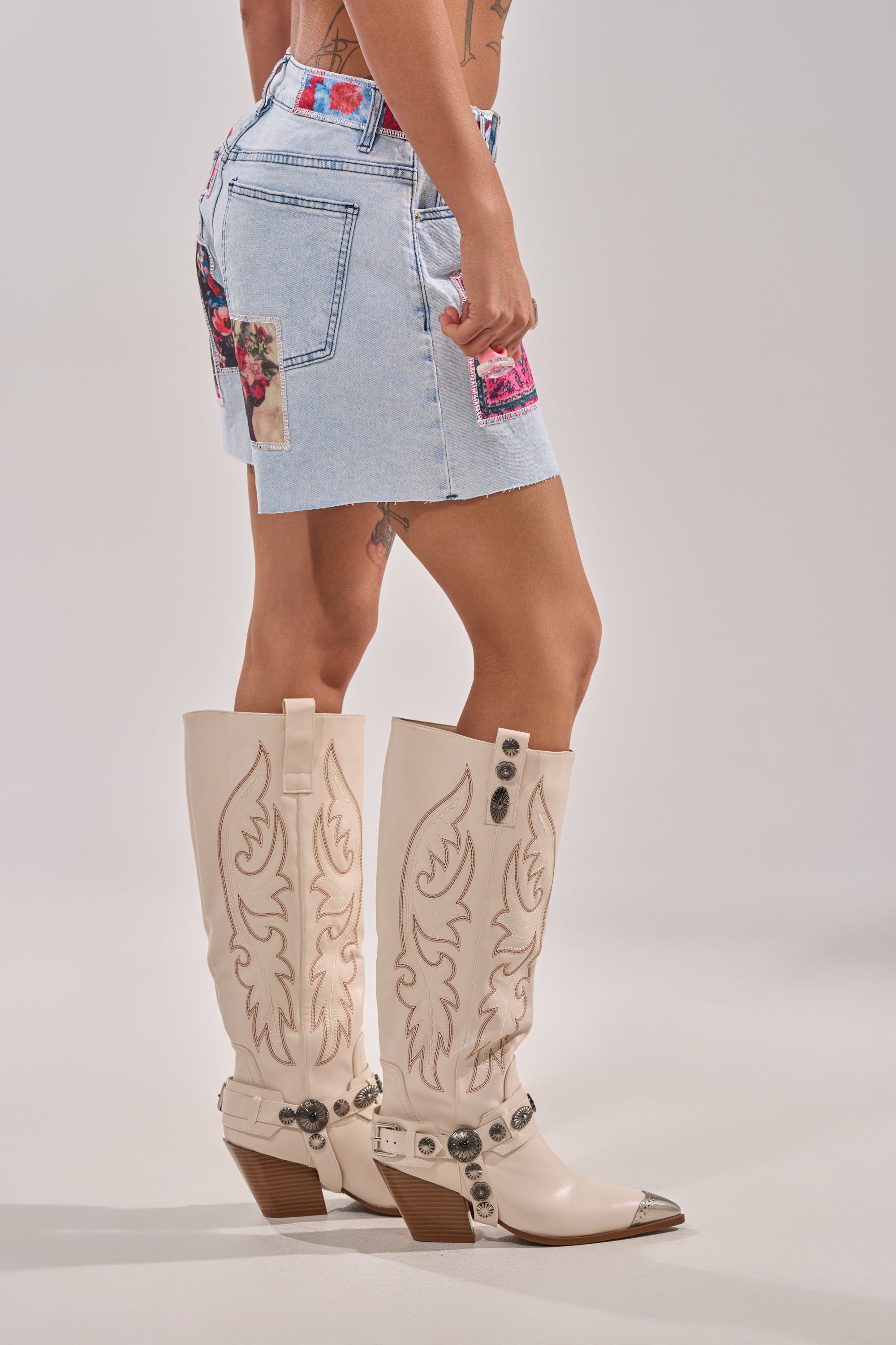 Wearing GET THE PICTURE PATCHWORK DENIM SHORTS, a light blue mini with colorful patches and high-rise waist, paired with beige knee-high cowboy boots with decorative stitching and metal accents against a plain background. 3