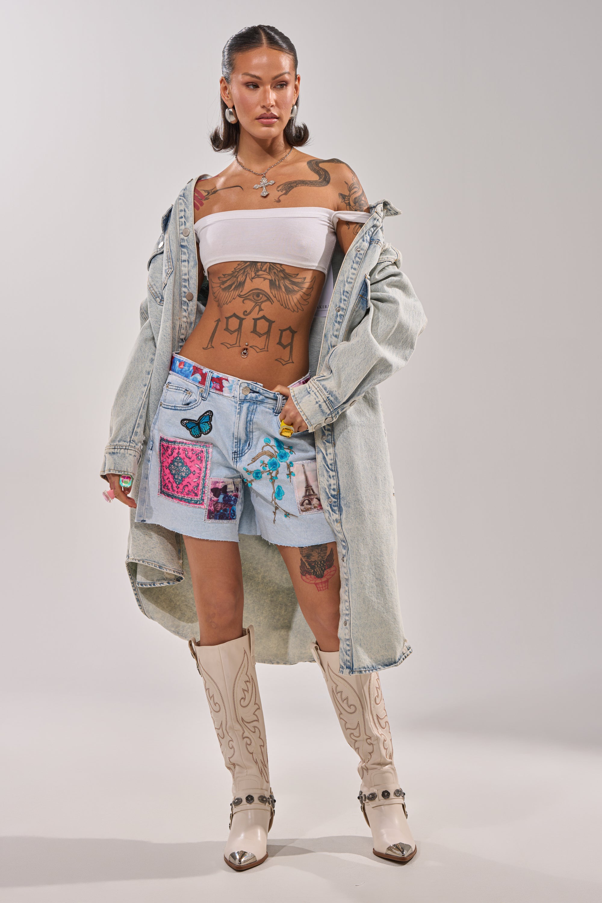A woman with tattoos models GET THE PICTURE PATCHWORK DENIM SHORTS with a raw-cut hem, paired with a white bandeau top, an oversized denim jacket, and cowboy boots as she poses confidently against a plain background. 6
