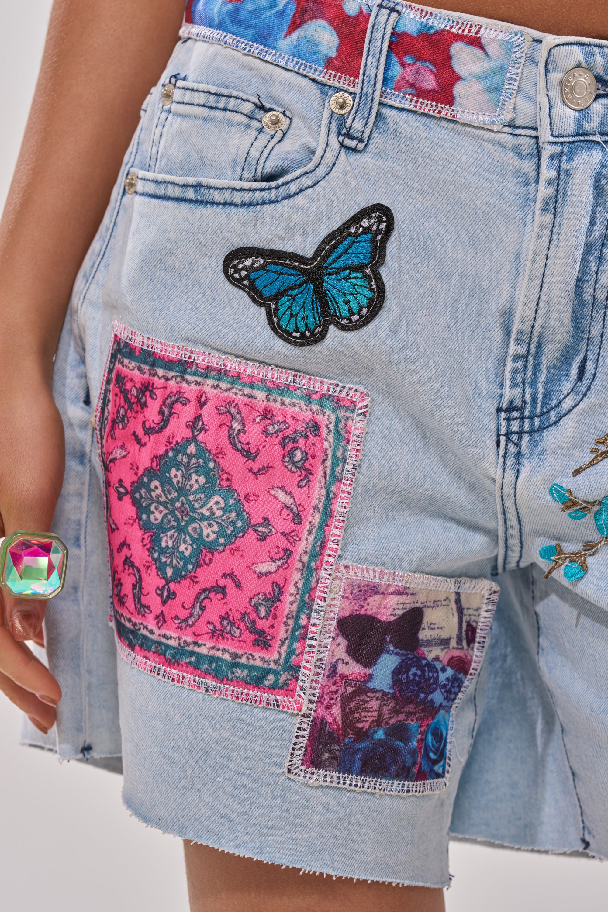 Close-up of a person in GET THE PICTURE PATCHWORK DENIM SHORTS, featuring a high-rise waist, raw-cut hemline, and colorful patches like a blue butterfly and photo print. They also wear a large faceted iridescent ring. 8