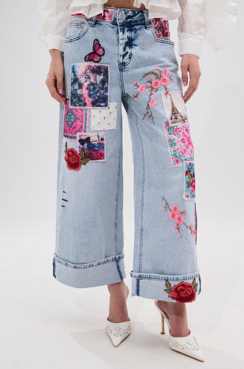 Someone models the PICTURE PERFECT PATCHWORK DENIM JEANS with colorful floral patches, styled with a white long-sleeve top and white heels, standing against a plain background.
