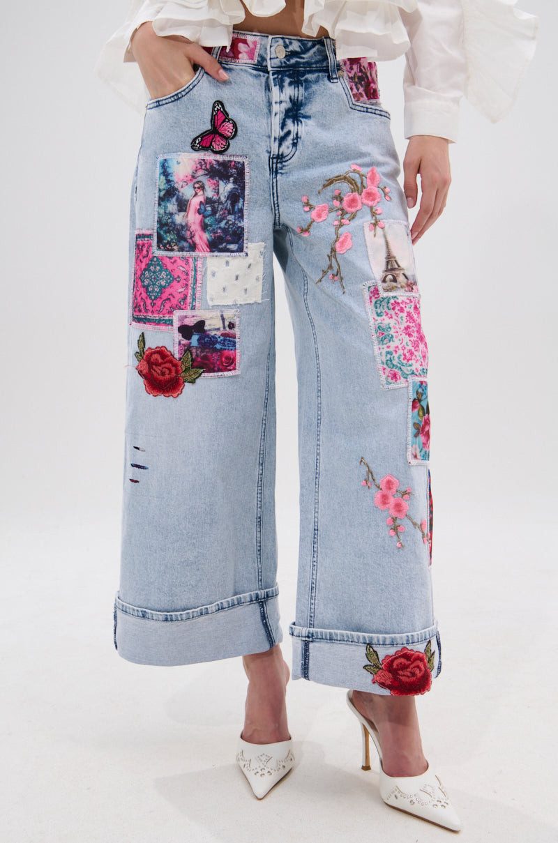 A person wears PICTURE PERFECT PATCHWORK DENIM JEANS featuring colorful patches and embroidery, paired with a white blouse and white heels.