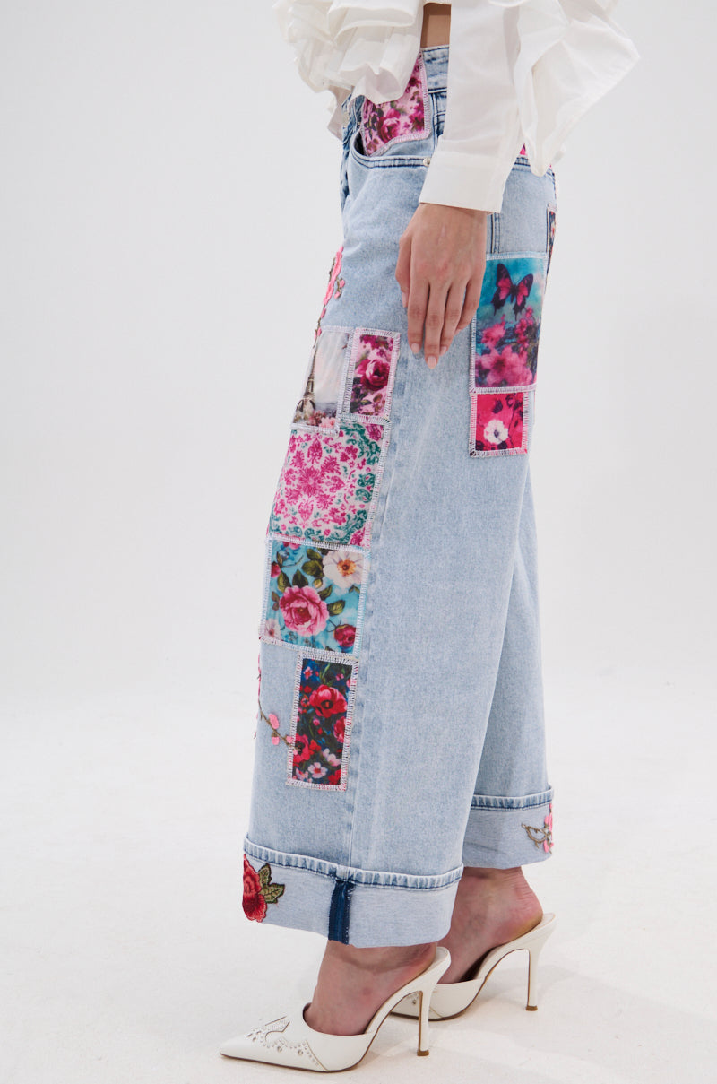 A person in PICTURE PERFECT PATCHWORK DENIM JEANS, a white ruffled blouse, and white pointed heels stands against a plain white background.