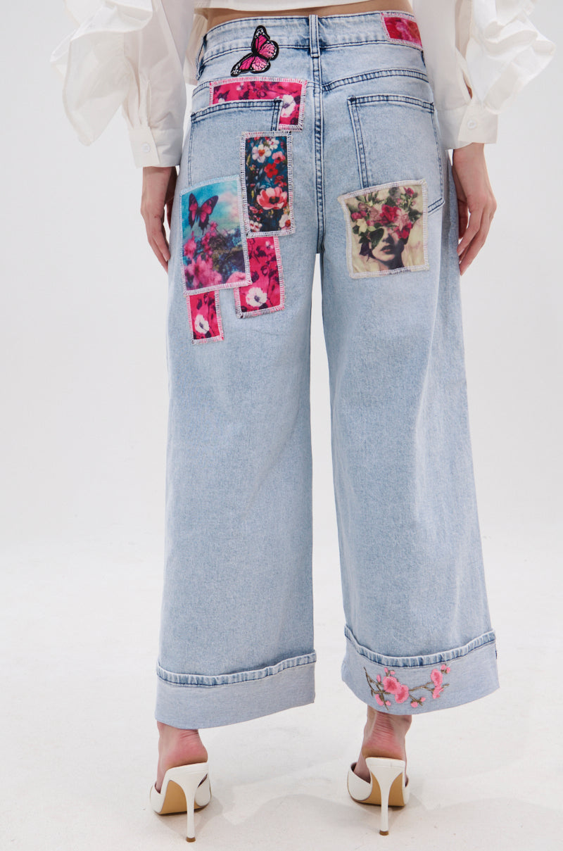 A person models the PICTURE PERFECT PATCHWORK DENIM JEANS, featuring high-rise wide legs and vibrant floral and butterfly patches on the back and hems, paired with white heels against a white background.