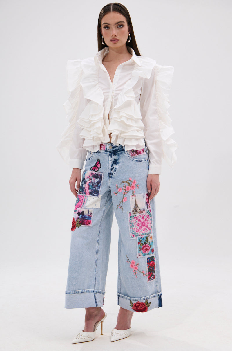 A woman stands against a white background wearing PICTURE PERFECT PATCHWORK DENIM JEANS—light blue high-rise wide-leg jeans with vibrant floral and butterfly accents—paired with a white ruffled blouse and white heels.
