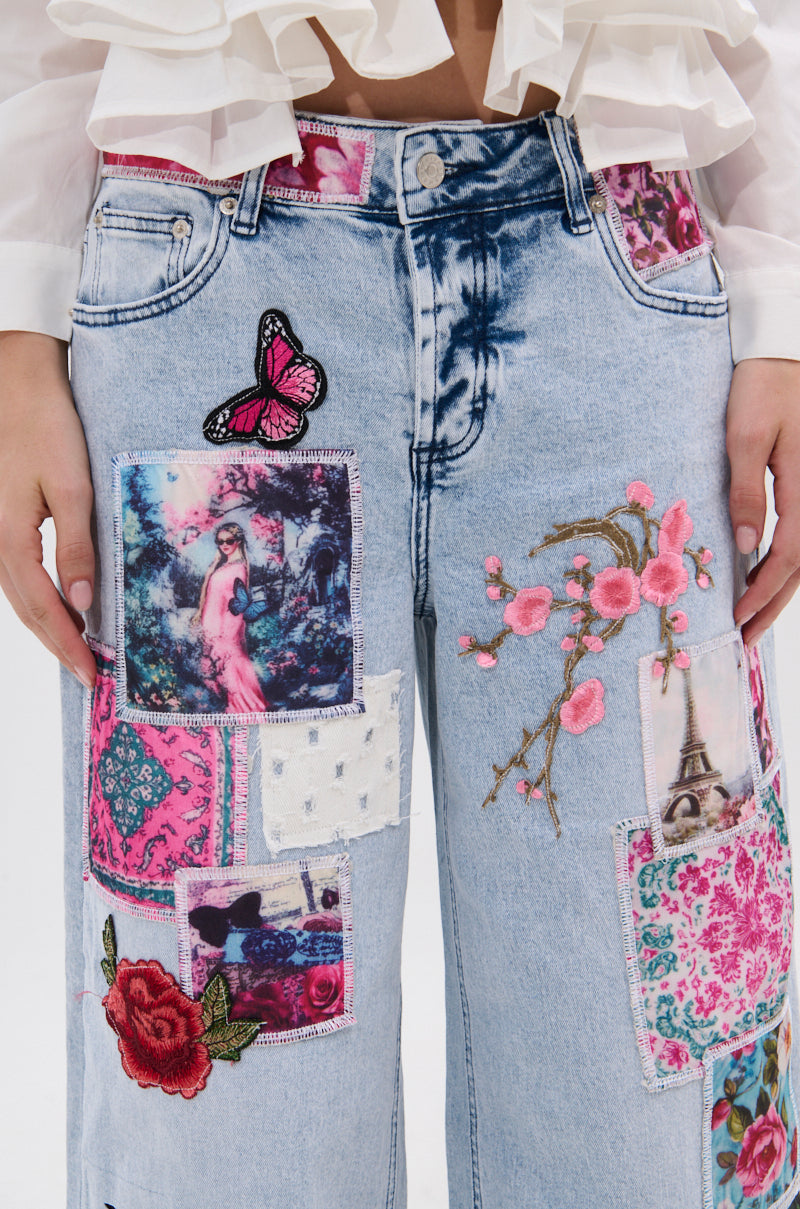 A close-up of someone in PICTURE PERFECT PATCHWORK DENIM JEANS, featuring colorful fabric patches, embroidered flowers, the Eiffel Tower, a butterfly, and vintage-style images. Part of their white ruffled shirt is also visible.