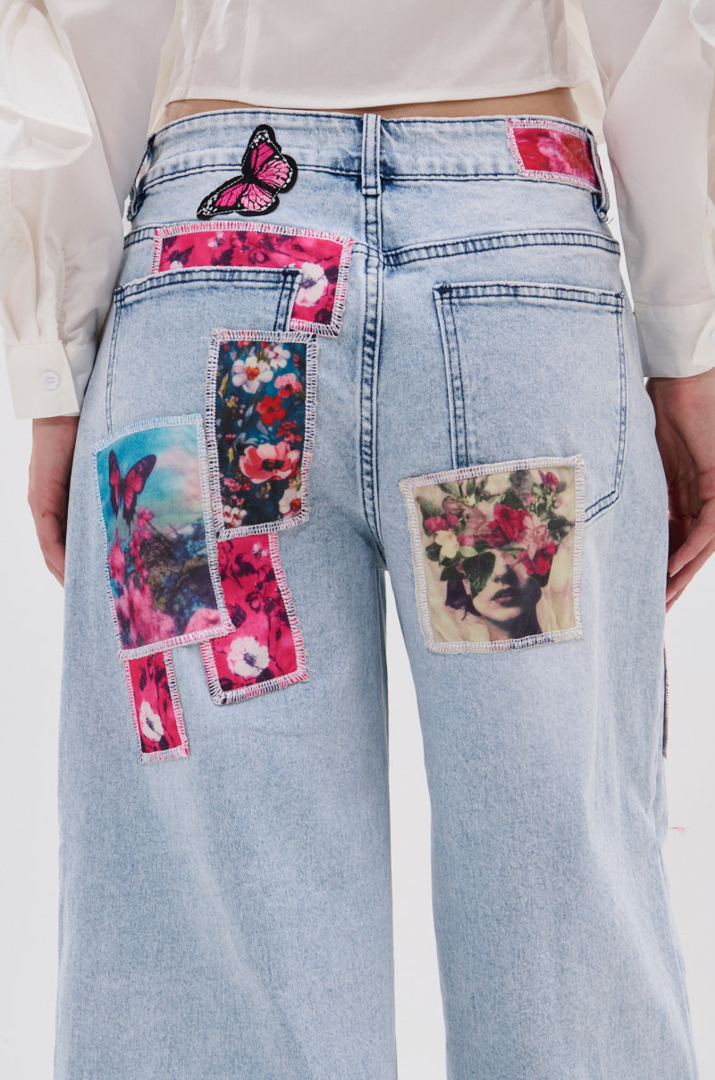 A person wears the PICTURE PERFECT PATCHWORK DENIM JEANS, featuring light blue denim with floral, butterfly, and face patchwork designs. The high-rise wide-leg jeans are paired with a white shirt visible above the waistband.