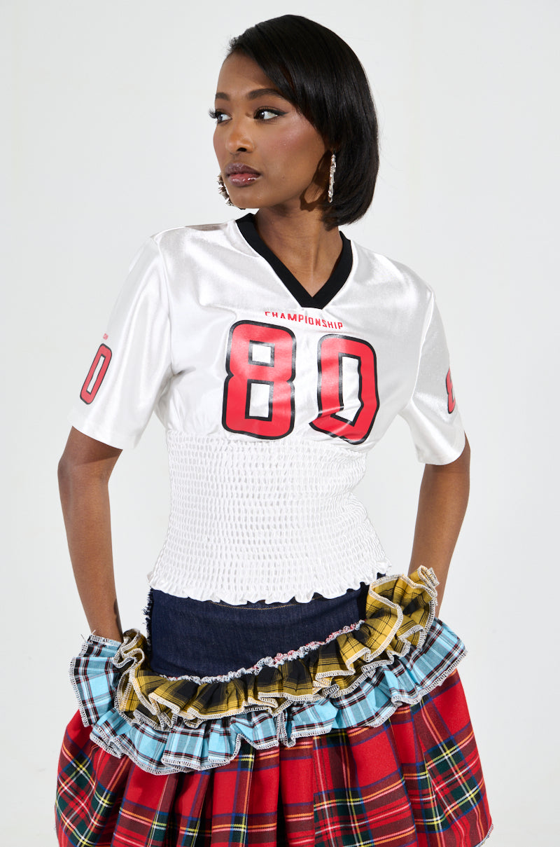 A woman wearing the sporty-chic "WANNA JOIN MY TEAM SCRUNCH JERSEY" top with the number 80, paired with a vibrant, layered skirt showcasing different plaid patterns. She has medium-length dark hair and is gazing to the side against a plain, light-colored background.