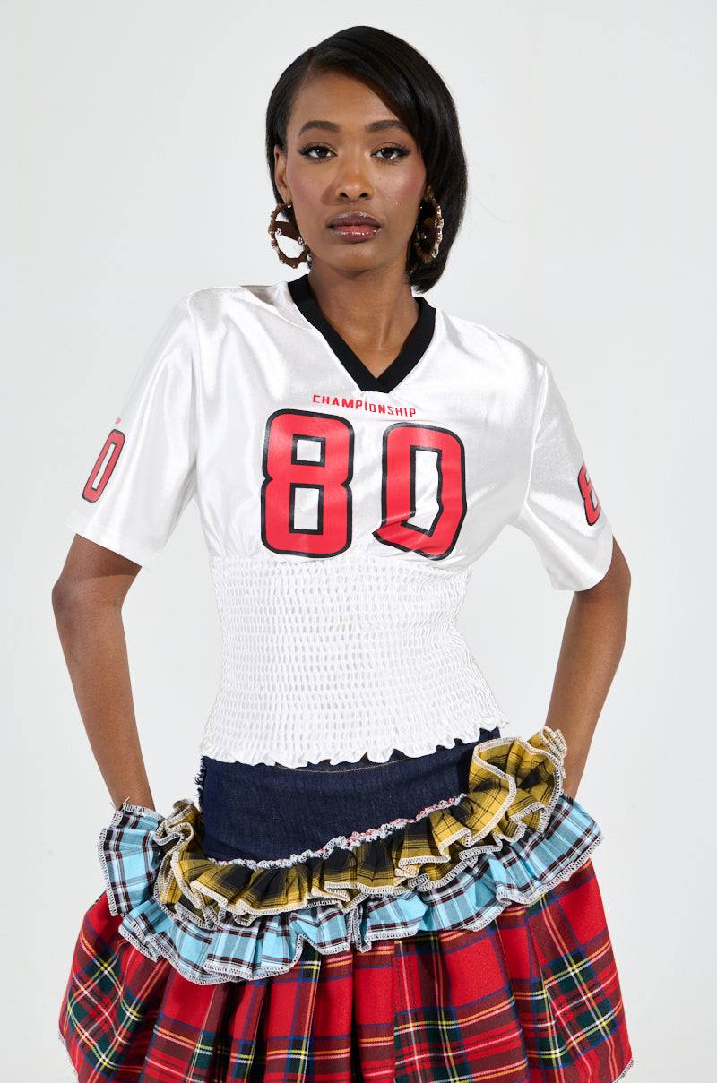 A woman stands confidently in a sporty-chic WANNA JOIN MY TEAM SCRUNCH JERSEY, adorned with Championship 80 graphics. The top is perfectly paired with her colorful layered plaid skirt and hoop earrings, while her neatly styled hair enhances the plain white background.