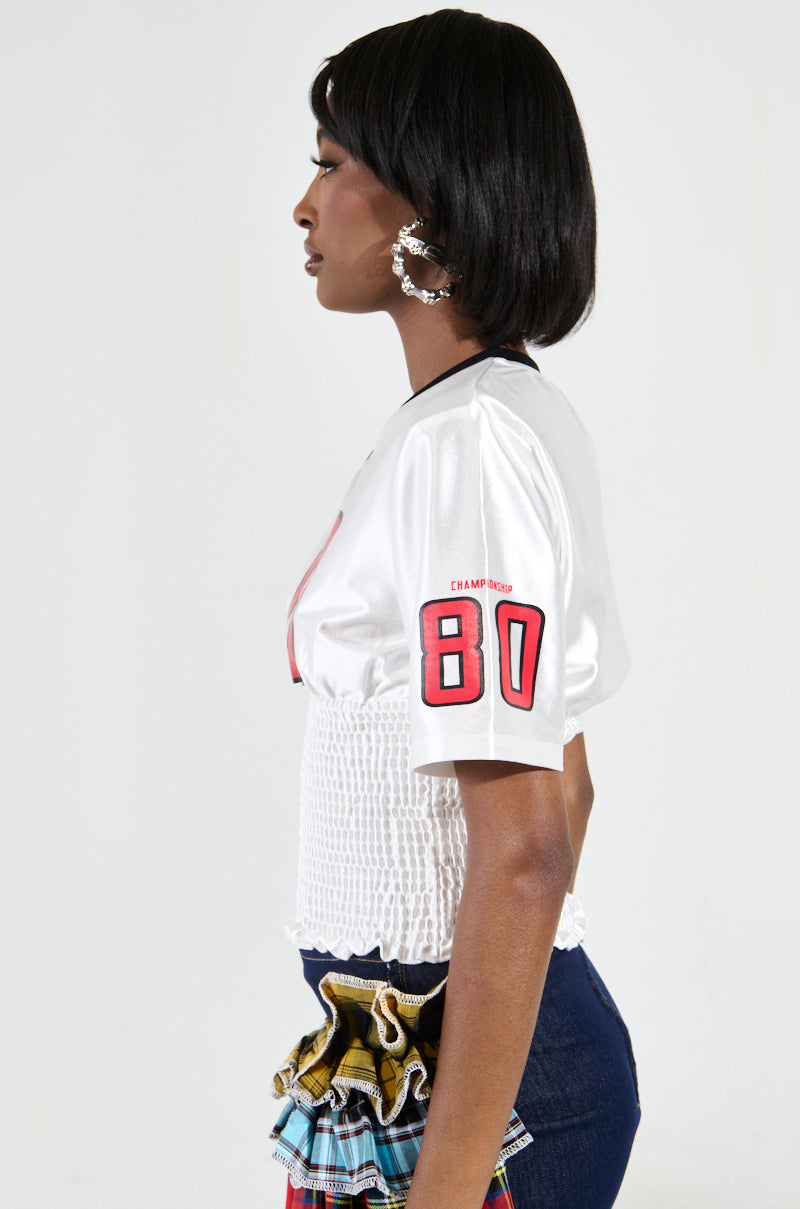 Sporting a short hairstyle, the individual models the WANNA JOIN MY TEAM SCRUNCH JERSEY with Championship 80 graphics on the sleeve. They pair this sporty-chic top with a smocked blouse and a denim skirt enriched by colorful ruffled details, completing the look with large hoop earrings. The scene captures their profile set against a plain backdrop.
