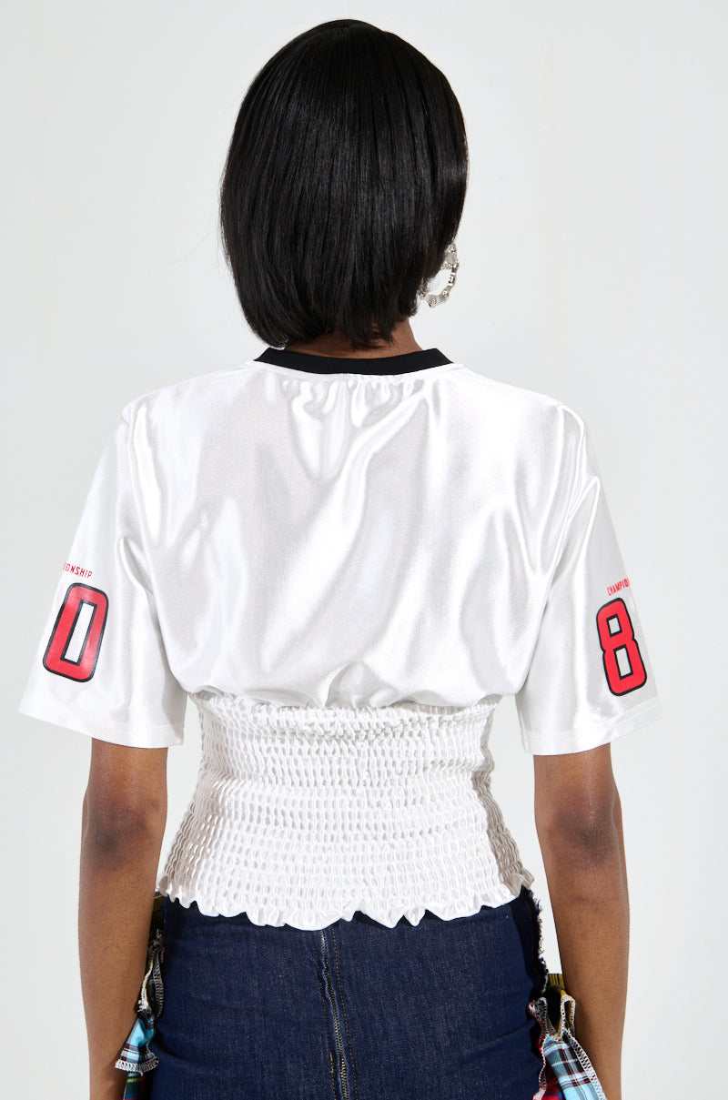 A person with short black hair is facing away, wearing the WANNA JOIN MY TEAM SCRUNCH JERSEY, a white sports jersey featuring the number 8 on the sleeves and a corset-style, sporty-chic design. They pair it with jeans adorned with decorative patches and a ruched waistband.