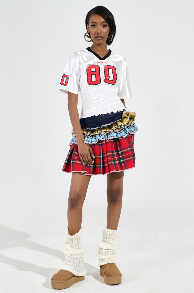 A person poses wearing the WANNA JOIN MY TEAM SCRUNCH JERSEY, featuring sporty-chic Championship 80 graphics. The outfit is complemented with a layered denim and red plaid skirt, tan boots, and thick cream socks against a plain white background.