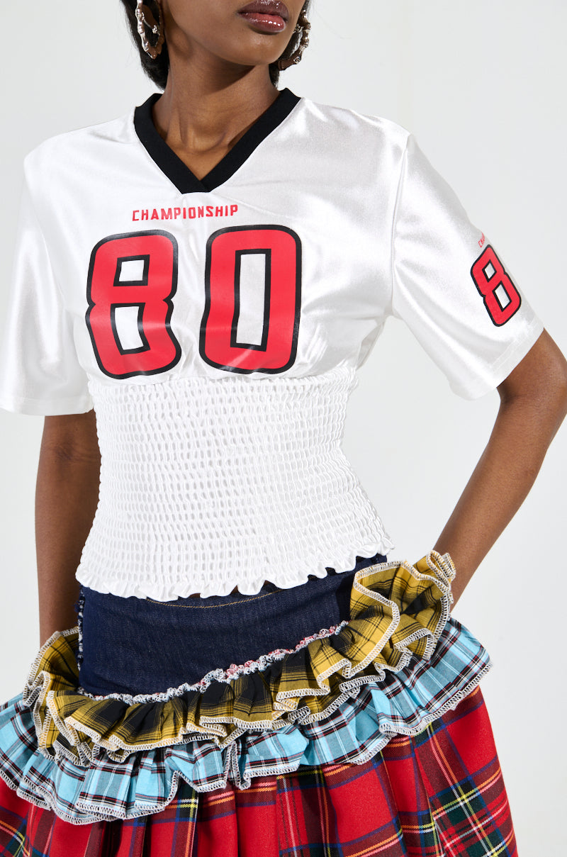 A person wearing the "WANNA JOIN MY TEAM SCRUNCH JERSEY" with Championship 80 graphics, paired with a colorful plaid skirt featuring ruffles and a ruched waistband. The outfit seamlessly blends sporty and eclectic styles.