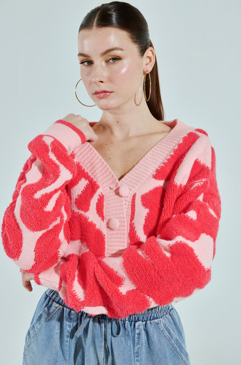 A person with long hair is wearing the AYANNA BUTTON UP CARDIGAN in fuzzy pink and red, featuring large buttons and a cropped silhouette. Their loose-fitting blue jeans complement the look, accessorized with large hoop earrings against a light gray background, arms crossed over their chest.