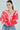 A person with long hair is wearing the AYANNA BUTTON UP CARDIGAN in fuzzy pink and red, featuring large buttons and a cropped silhouette. Their loose-fitting blue jeans complement the look, accessorized with large hoop earrings against a light gray background, arms crossed over their chest.
