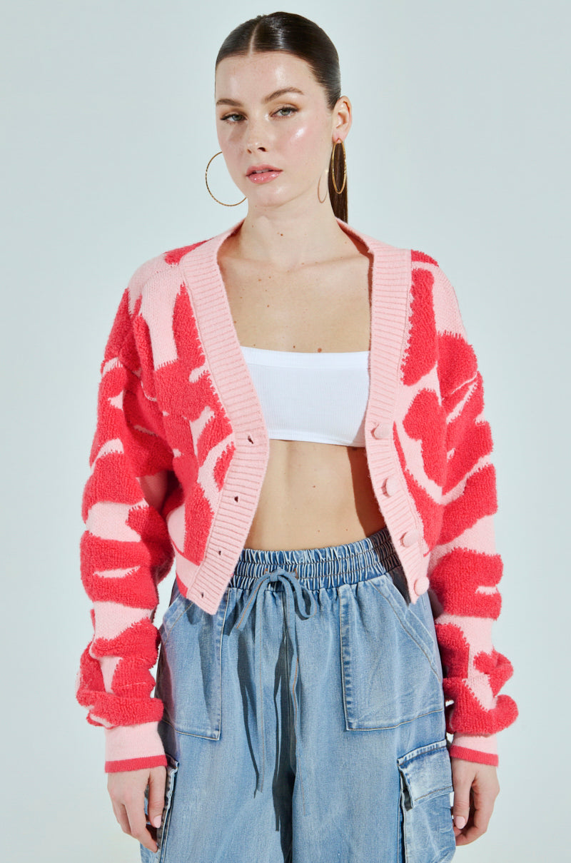 A person stands against a plain background wearing the AYANNA BUTTON UP CARDIGAN, featuring a pink and red pattern with a V-neckline, paired with a white crop top and blue drawstring pants. They have long hair pulled back and accessorize with large hoop earrings.