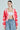 A person stands against a plain background wearing the AYANNA BUTTON UP CARDIGAN, featuring a pink and red pattern with a V-neckline, paired with a white crop top and blue drawstring pants. They have long hair pulled back and accessorize with large hoop earrings.
