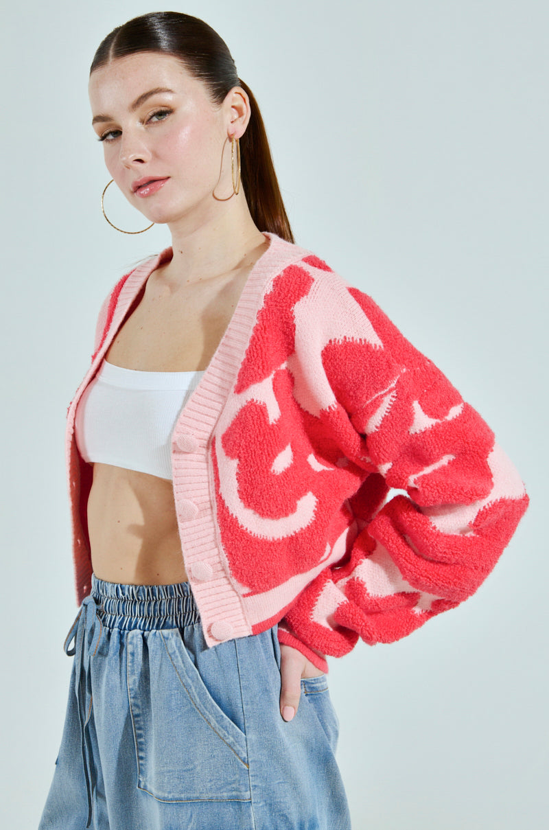 A person with long brown hair wears the AYANNA BUTTON UP CARDIGAN in pink and red, featuring a V-neckline, over a white cropped top and loose blue jeans. They sport large hoop earrings and have one hand in their pocket, standing against a plain background.