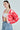 A person with long brown hair wears the AYANNA BUTTON UP CARDIGAN in pink and red, featuring a V-neckline, over a white cropped top and loose blue jeans. They sport large hoop earrings and have one hand in their pocket, standing against a plain background.