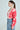 A woman with a ponytail wears large hoop earrings and the AYANNA BUTTON UP CARDIGAN, featuring a pink and red pattern with a V-neckline over a white top. Her high-waisted blue pants enhance the cropped silhouette as she stands in profile against a plain background.
