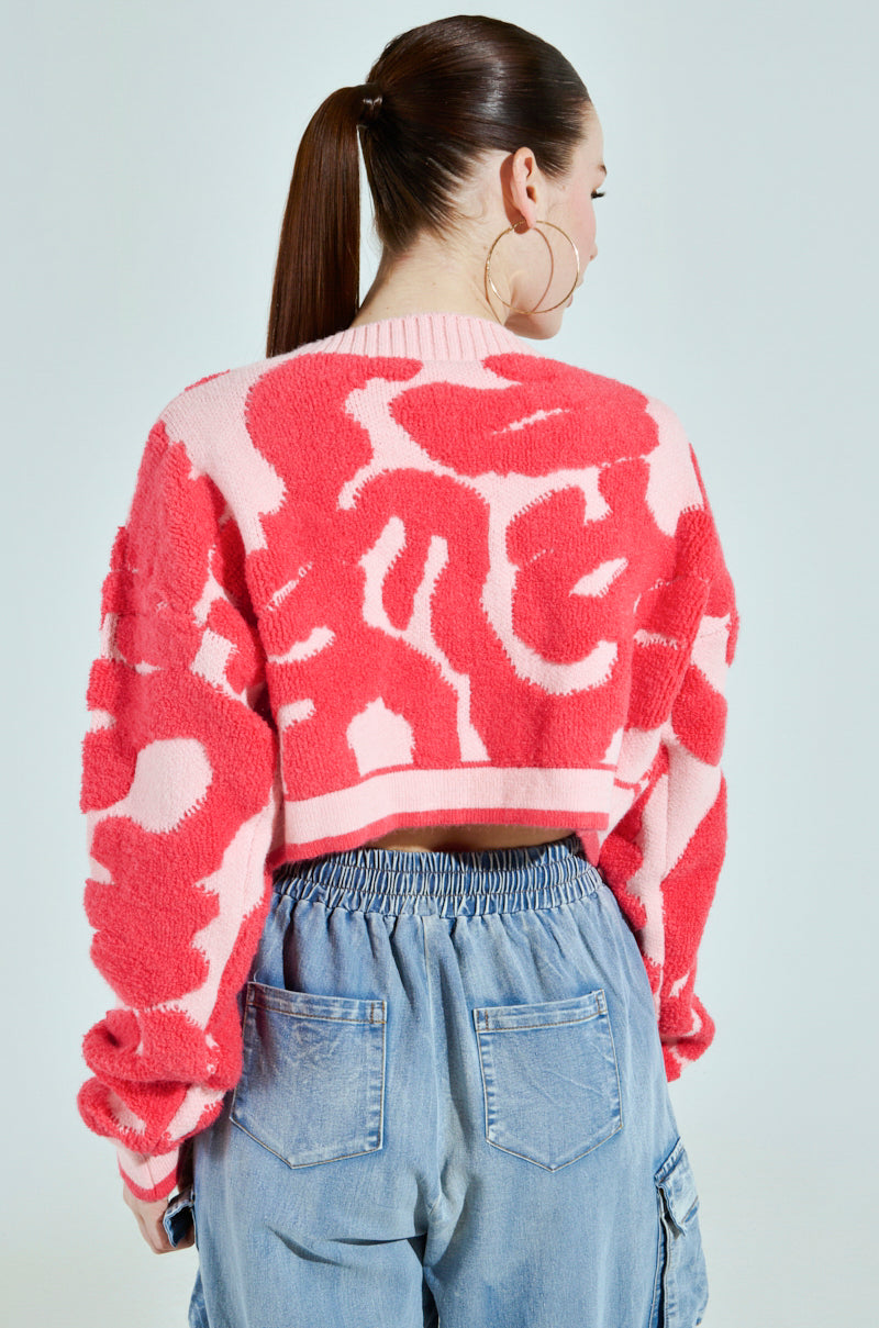 The person with a ponytail wears the AYANNA BUTTON UP CARDIGAN, a large pink and red patterned V-neck sweater, paired with loose-fitting blue jeans featuring an elastic waistband. They are facing away from the camera against a light background.