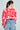 The person with a ponytail wears the AYANNA BUTTON UP CARDIGAN, a large pink and red patterned V-neck sweater, paired with loose-fitting blue jeans featuring an elastic waistband. They are facing away from the camera against a light background.