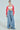 A person models the AYANNA BUTTON UP CARDIGAN, a pink patterned V-neck design, over a white tube top, with wide-leg denim cargo pants and beige shoes. Their hair is slicked back, complemented by large hoop earrings against a light-colored background.