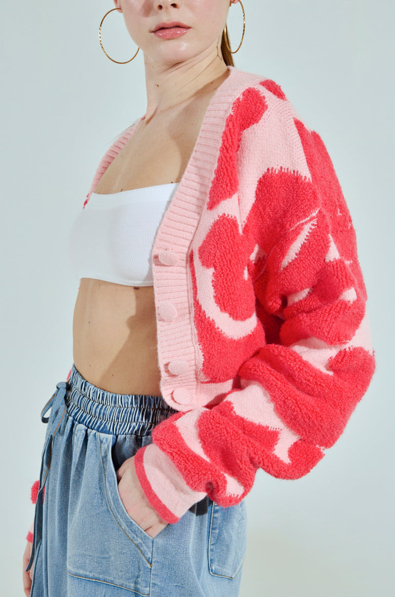 A person wears the AYANNA BUTTON UP CARDIGAN in a pink-red pattern with a V-neck over a white bandeau top, paired with light blue wide-legged jeans and large gold hoop earrings, creating a stylish cropped silhouette against a plain white background. One hand rests casually in their pocket.