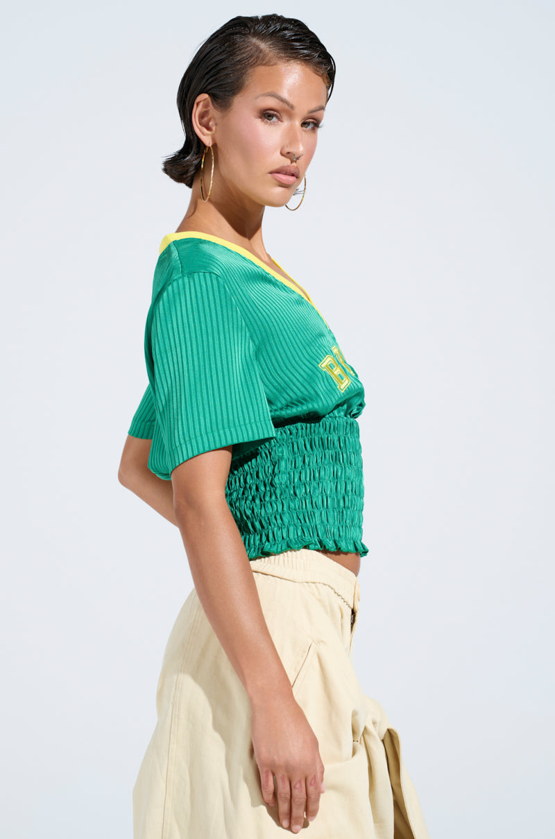 A woman with slicked-back hair exudes sporty chic style in large hoop earrings and a BRASIL RUCHED JERSEY, accessorized with beige pants. She poses confidently against a light blue background, her gaze fixed intently on the camera.