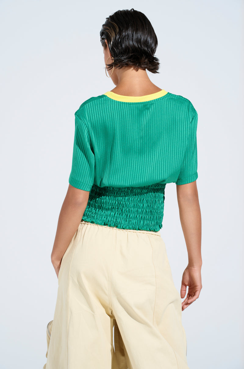 A person with short dark hair faces away, wearing a BRASIL RUCHED JERSEY with a yellow neckline and cream wide-leg pants, set against a light background.