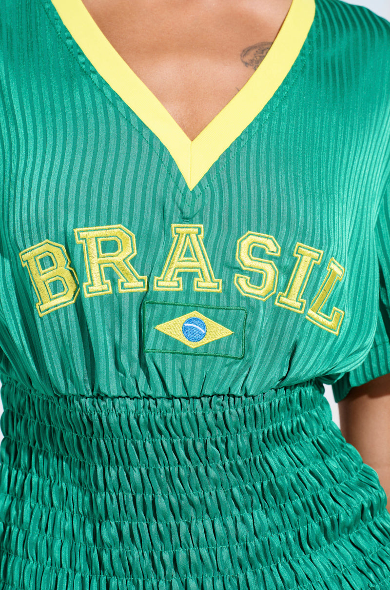Close-up of the BRASIL RUCHED JERSEY: a green jersey with a yellow v-neck, blending sporty chic and flair. It features "Brasil" lettering and a small Brazilian flag emblem, highlighting its textured, ruched design below the chest.