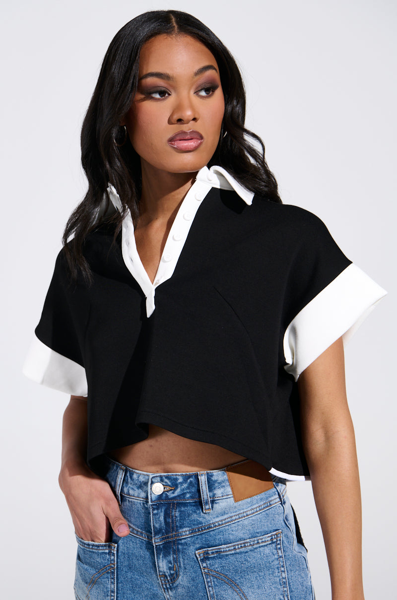 A person with long black hair confidently poses against a plain background, hand on hip, wearing blue jeans and the stylish NOTHING TO SEE BLOUSE by AKIRA Label. The blouse features a white collar and cuffs, is slightly cropped, revealing a hint of their lower back. 1