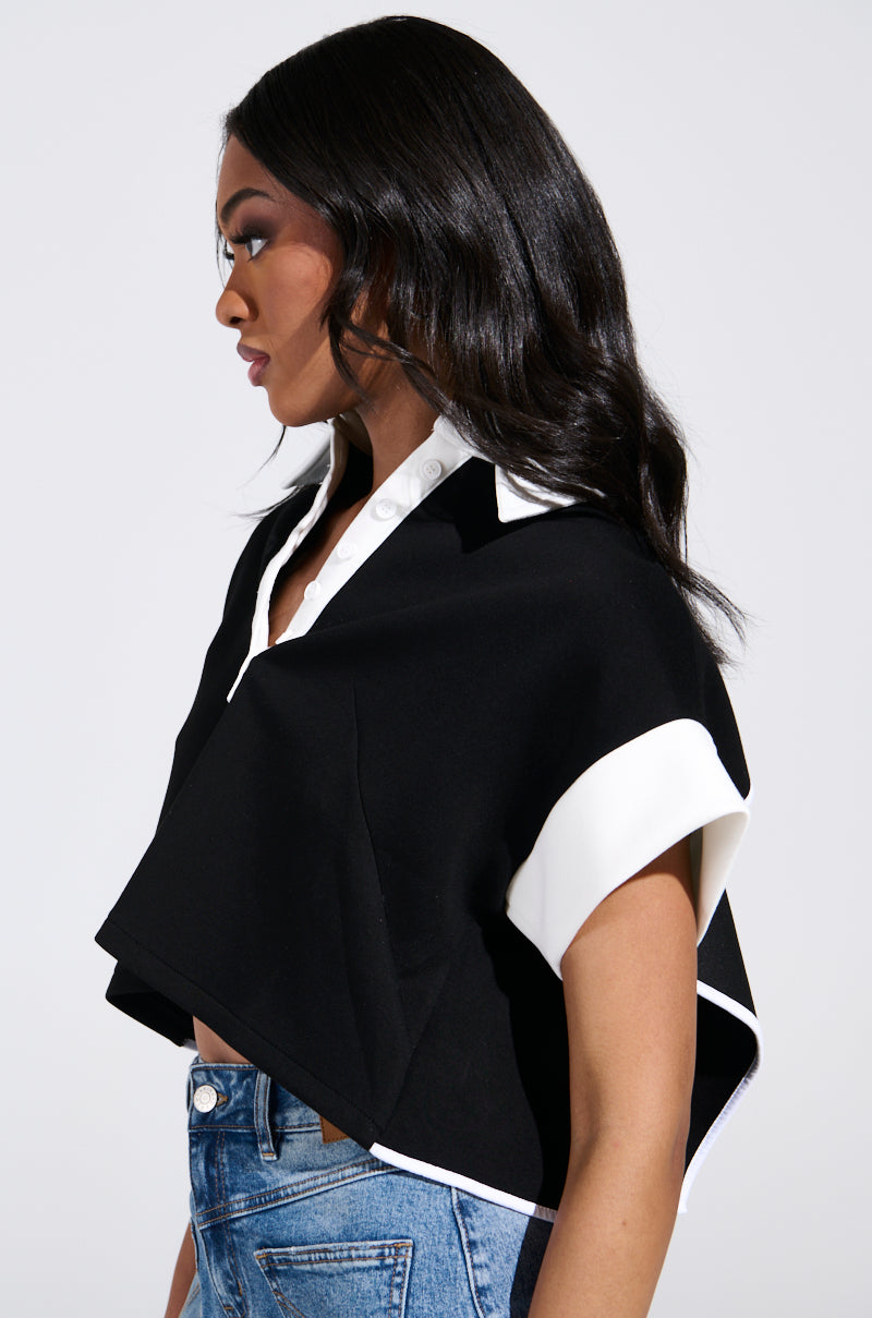 A side view shows a woman with long dark hair in the NOTHING TO SEE BLOUSE. This black and white stretchy cropped top features a collared neckline and short sleeves, exposing her lower back. Her high-waisted blue jeans pair well with the top as she stands against a plain background. 2
