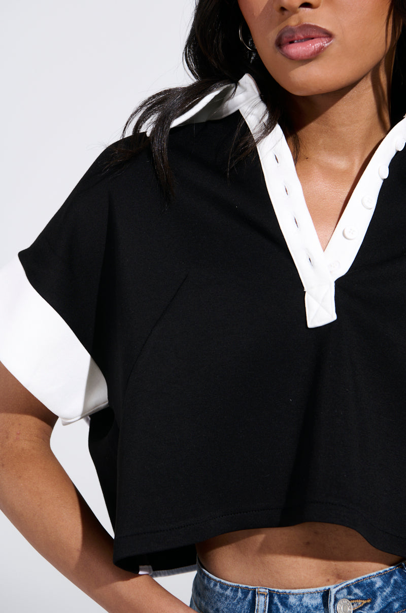 A person is wearing the AKIRA Label NOTHING TO SEE BLOUSE in black and white. This stretchy, cropped top has button details, short sleeves, and an exposed lower back. They pair it with blue denim jeans against a plain, light background. 7