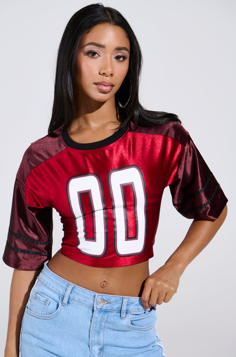 A woman with long dark hair striking a pose against a plain background wears the sporty-chic DOUBLE TROUBLE JERSEY, which is red and cropped with a lace-up panel featuring the number "00," paired with light blue jeans.