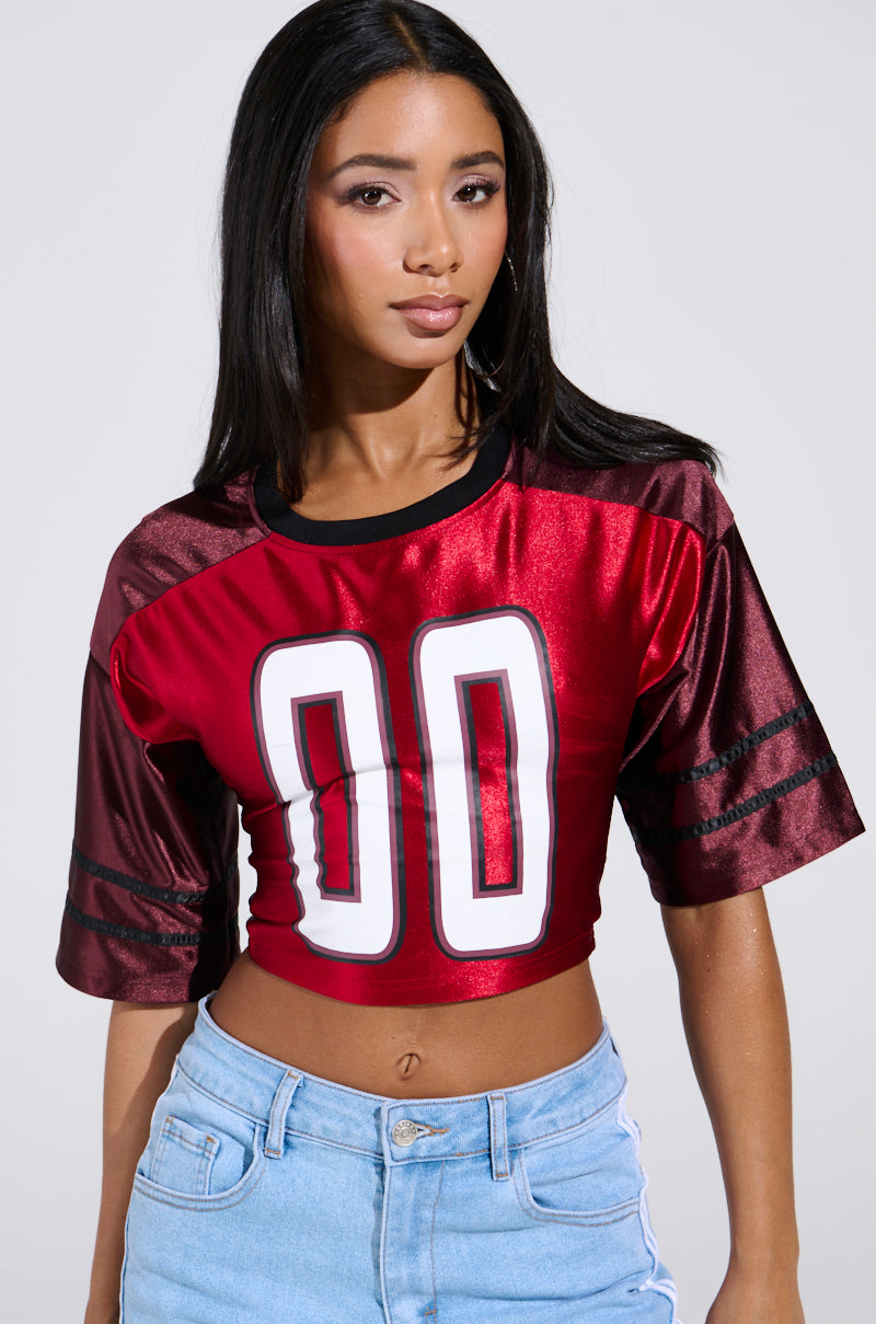 Sporting long black hair, a person wears the DOUBLE TROUBLE JERSEY crop top in shiny red and maroon with "00" on it, paired with light blue jeans and featuring black trim on the sleeves against a plain, neutral background.