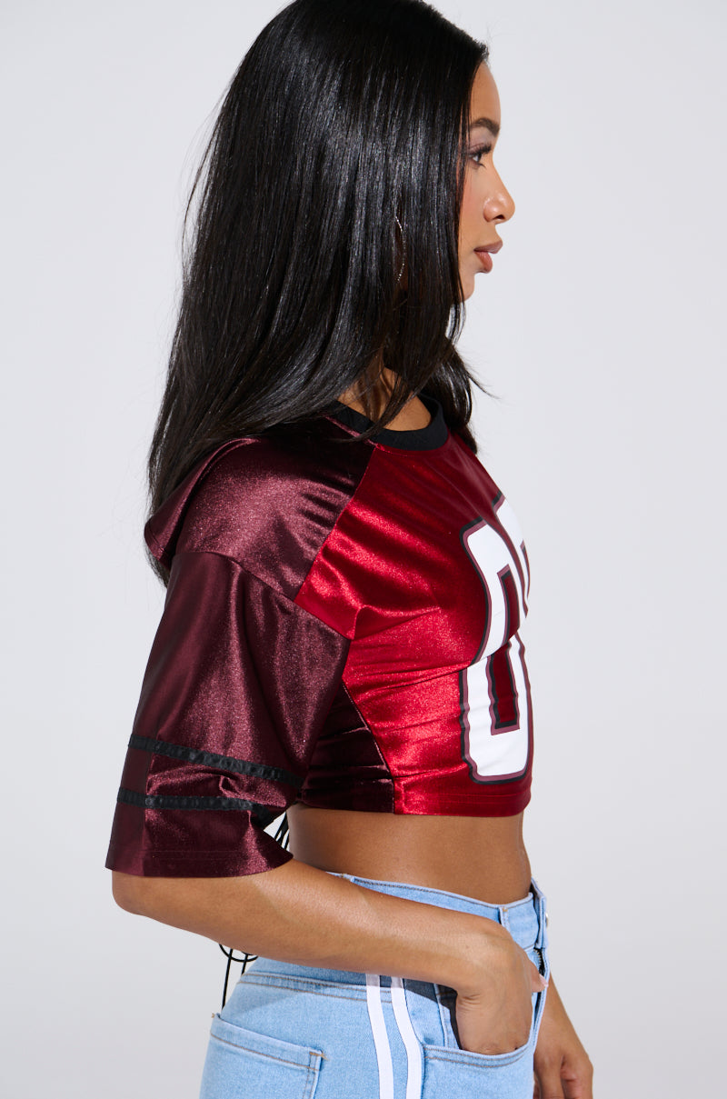A woman with long black hair sports the DOUBLE TROUBLE JERSEY, a sporty-chic maroon crop top featuring large white numbers in a color-block design, paired with light blue jeans. She is shown in profile against a plain white background.