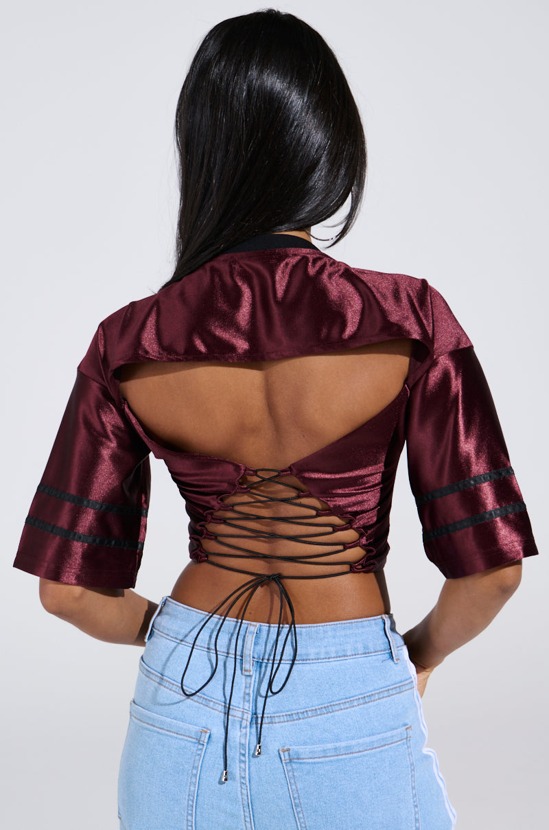 Rear view of a person with long dark hair wearing the DOUBLE TROUBLE JERSEY, a satin burgundy sporty-chic top featuring lace-up back and open cut-out design, paired with light blue jeans.