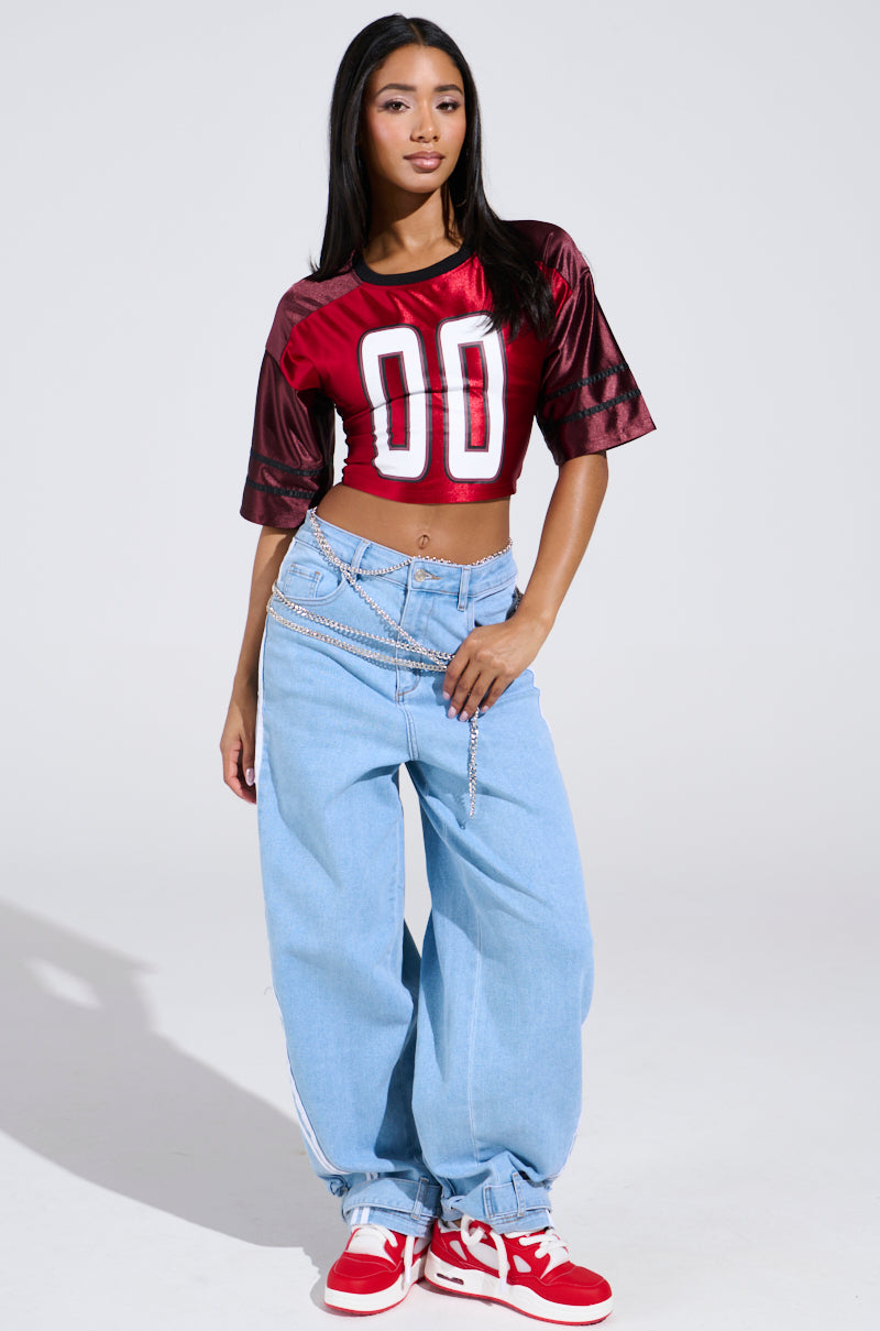 A woman confidently poses in a sporty-chic red DOUBLE TROUBLE JERSEY with "00," light blue baggy jeans, red and white lace-up sneakers, and a chain belt against a plain background.