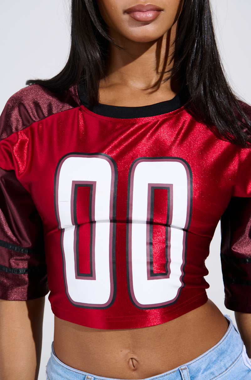 A woman wears a Double Trouble Jersey, a shiny red sporty-chic crop top with "00" on the front, paired with light blue jeans. Her long dark hair falls over her shoulders, enhancing her neutral expression against a minimalist white background that highlights the sleek color-block design.