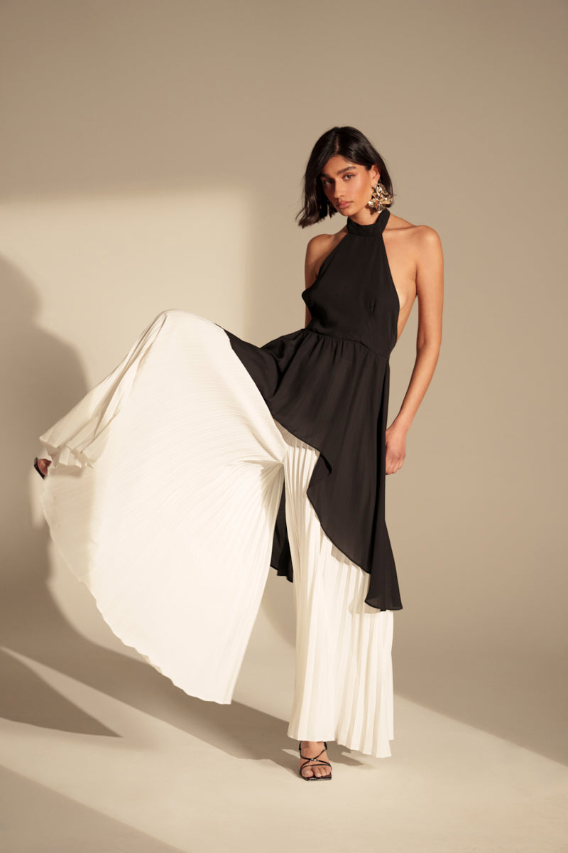A woman showcases the FLY INTO THE AIR PALAZZO PANT with a sleeveless black halter top. The wide-leg pants flow as she lifts one leg. Strappy heels and statement earrings finish her style, set against a neutral backdrop.