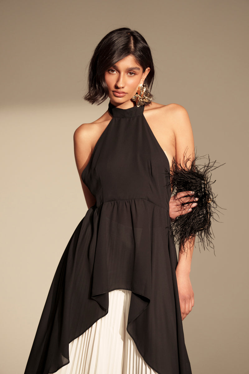 A woman with a bob haircut wears the FLY INTO THE AIR BLOUSE, a sleeveless black chiffon high-neck halter top with an uneven hem, styled over a white pleated skirt. She holds a black feathered accessory and wears statement earrings against a beige background.