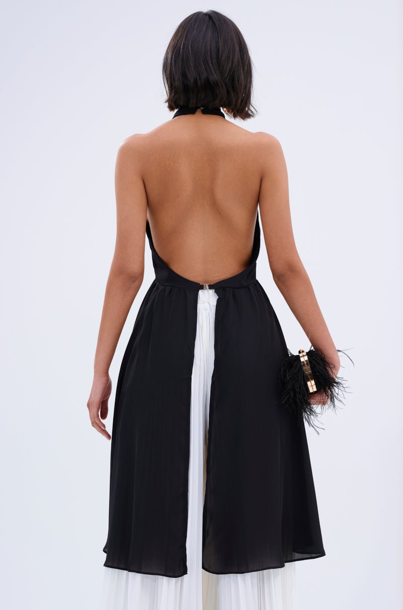 A woman with short dark hair is shown from behind wearing the FLY INTO THE AIR BLOUSE, a sleeveless high-neck top, paired with a black dress over a white skirt. She holds a small black feather-detailed clutch against a plain light background.