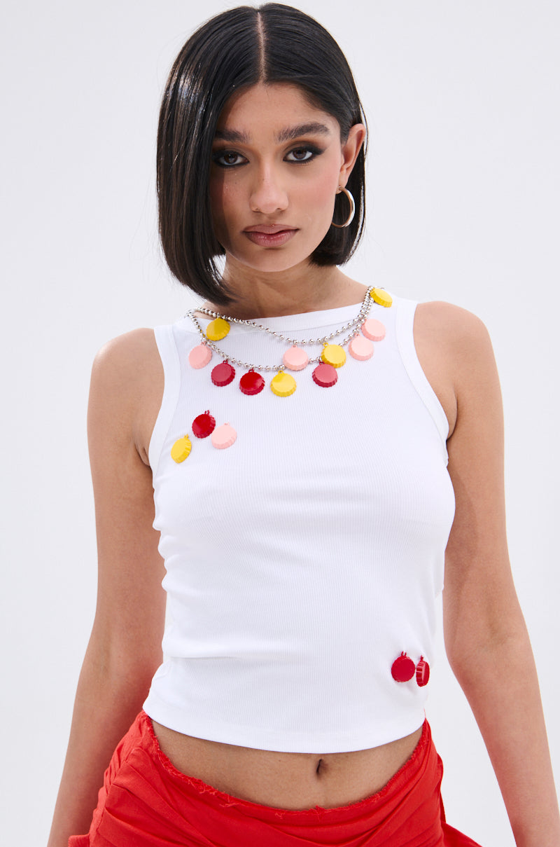 A woman with straight black hair models the GIRL POWER TANK—a sleeveless white top with a scoop neckline and colorful circular accents—styled with a chunky necklace and red wrap skirt against a plain light background.