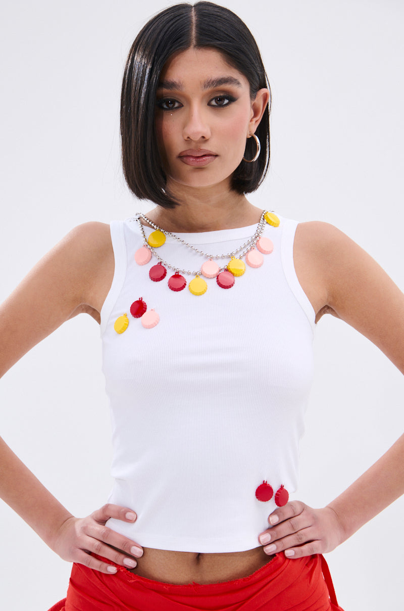 A woman with a sleek bob wears the GIRL POWER TANK, a white ribbed knit top with a scoop neckline, paired with red pants and a colorful charm necklace. She stands confidently with hands on hips against a plain background.